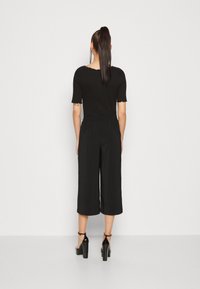 Pantalon noir court avec une ceinture élastique, associé à un haut noir à manches courtes avec un ourlet festonné. Le mannequin porte des talons hauts noirs.