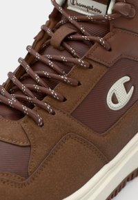 Brown high-top sneakers with textured suede and mesh panels, featuring brown laces with white specks and a logo emblem on the side.