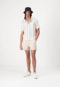 Korte mouw knopenhemd met verticale strepen, beige shorts en witte sneakers. De persoon draagt een zwarte bucket hat. Eenvoudige texturen.