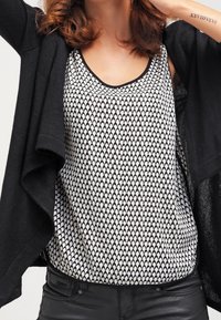 Gilet noir ouvert sur un haut sans manches blanc avec un motif en écailles noires. Un pantalon noir ajusté est visible en bas. Tissu texturé.