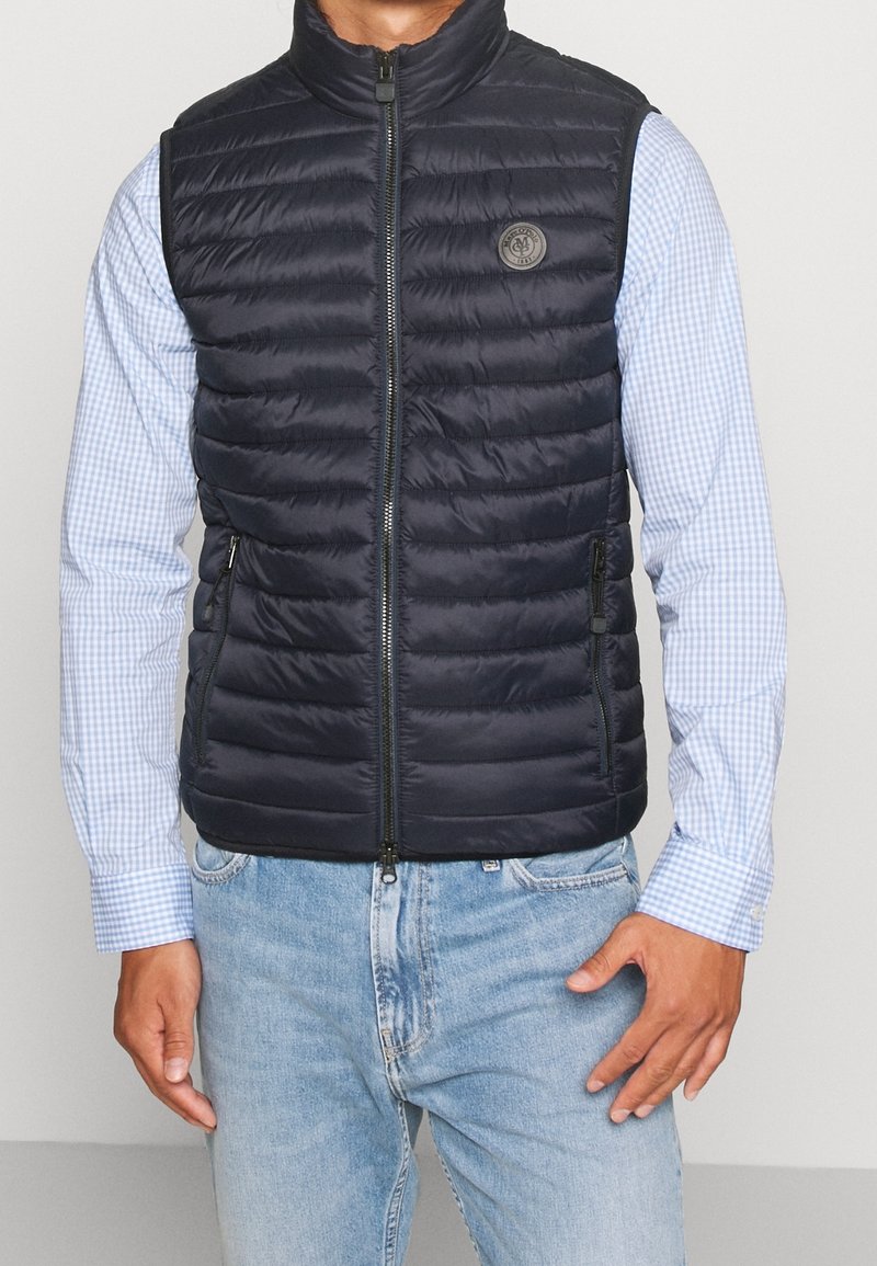Gilet imbottito blu navy con colletto alto, texture a trapunta orizzontale, zip frontale e tasche laterali. Indossato sopra una camicia a quadri blu chiaro.