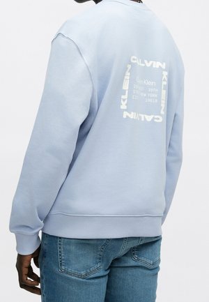 Personne portant un sweat-shirt Calvin Klein bleu clair avec un logo carré blanc et un texte au dos, associé à un jean bleu.
