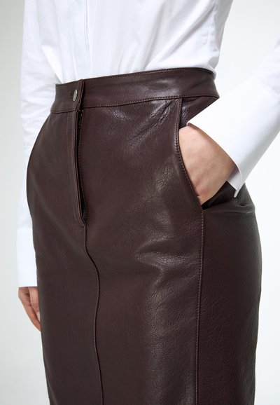 Detail osoby nosiacej tmavohnedú koženú sukňu s predným zipsom a vreckom, v kombinácii s bielou košeľou s dlhými rukávmi.