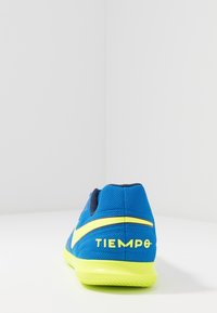 Zapatilla deportiva azul y amarillo neón con la marca "TIEMPO" en el talón, vista desde atrás sobre una superficie blanca.