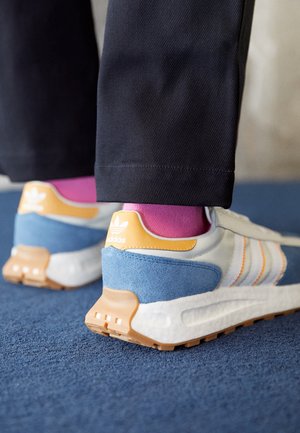 Personne portant des baskets blanches Adidas avec des détails bleus, orange et beiges, des chaussettes roses et un pantalon noir, debout sur un tapis bleu.