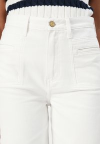 White denim high-waisted jeans feature a classic five-pocket design and a metal button closure, showcasing a smooth, textured fabric.
