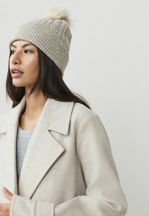 Beige stickad mössa med ett texturerat mönster, försedd med en fluffig pom-pom överst. Bärs med en mjuk grå kappa och en ribbad tröja.