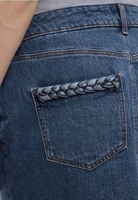 Πίσω τσέπη τζιν με πλεκτή λεπτομέρεια από λωρίδα τζιν κατά μήκος της πάνω άκρης, ορατή ραφή και μέρος του χεριού ενός ατόμου κοντά.