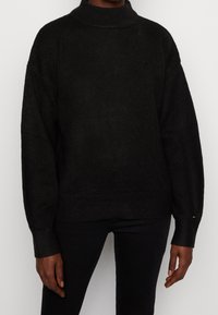 Pull noir en maille avec un col montant, coupe ample et épaules tombantes. Comprend des poignets et un ourlet côtelés, texture douce, branding minimaliste.
