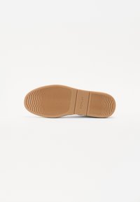 Suola della scarpa progettata in gomma con un motivo a onda, di colore beige e con branding in rilievo sulla parte centrale. Texture complessivamente liscia.