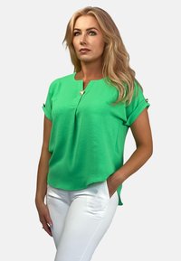 Zielony krótki rękaw w lekkim materiale z okrągłym dekoltem, ozdobiony detalem w postaci guzika, o luźnym kroju. W zestawie z białymi spodniami.