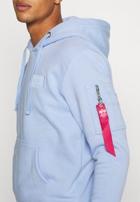 Felpa leggera blu chiaro realizzata in un tessuto morbido. Presenta una tasca frontale, cappuccio con cordini e una tasca con zip sul braccio con un'etichetta rossa.