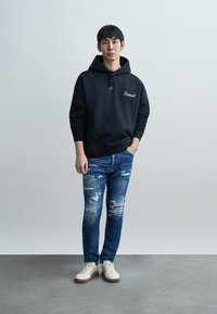 Μαύρο φούτερ με κουκούλα και λευκό λογότυπο, σε συνδυασμό με ανοιχτόμπλε τζιν με φθορές και λευκά αθλητικά παπούτσια με καφέ σόλες.