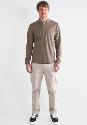 Polo shirt olive à manches longues avec col, associé à un pantalon beige clair et des baskets noires. Présente un petit logo sur la poitrine.