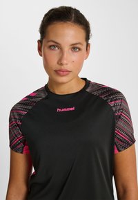 Koszulka sportowa z krótkim rękawem, z czarnym, jednolitym korpusem oraz różowo-szarymi wzorzystymi akcentami na ramionach i logo "hummel" w kolorze różowym.