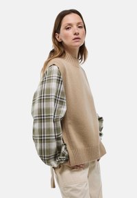 Pull en tricot sans manches beige avec un col montant superposé sur une chemise à carreaux verte à manches bouffantes. Pantalons de couleur crème visibles.