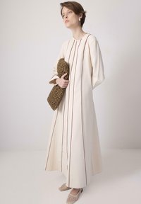 A woman stands elegantly in a long cream dress with brown stripes, holding a woven clutch, wearing stylish shoes, against a minimal backdrop.