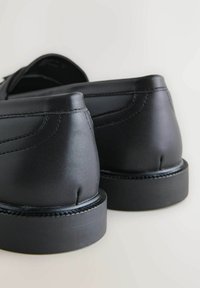 Czarne skórzane buty o gładkiej fakturze, zaokrąglonym obcasie i widocznym przeszyciu wzdłuż górnej krawędzi, umieszczone na jasnym tle.