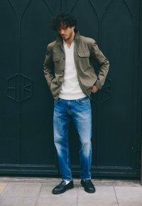 Olivza zaļā jaka ar divām priekšējām kabatām, balts džemperis, gaiši zili džinsi un melnas mokasīnes. Cirtaini mati un stāvoša poza tumšā fona priekšā.