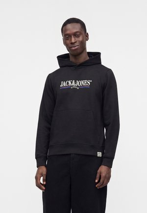 Homme portant un sweat à capuche noir Jack & Jones et un pantalon noir, debout sur un fond gris clair uni.