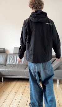 Czarna lekka kurtka z kapturem, z logo na plecach. Nosi się ją z luźnymi niebieskimi jeansami, na drewnianej podłodze obok szarej sofy.