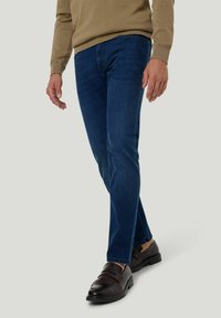 Jean en denim bleu foncé avec une coupe slim, présentant un design classique à cinq poches et un léger délavage, associés à des chaussures en cuir foncé à enfiler.