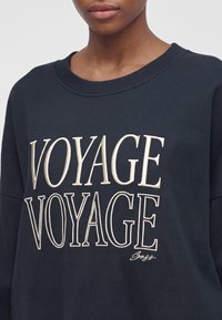 Човек, носещ тъмна блуза със думата "VOYAGE" написана два пъти с големи букви отпред.