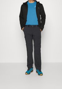 Man med svart zip-up jacka, blå grafisk t-shirt, mörka cargobyxor och blå trailrunning-skor som står med händerna i fickorna.