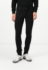 Black denim jeans with a slim fit, five-pocket design, and button closure. Paired with gray and white sneakers featuring black stripes.