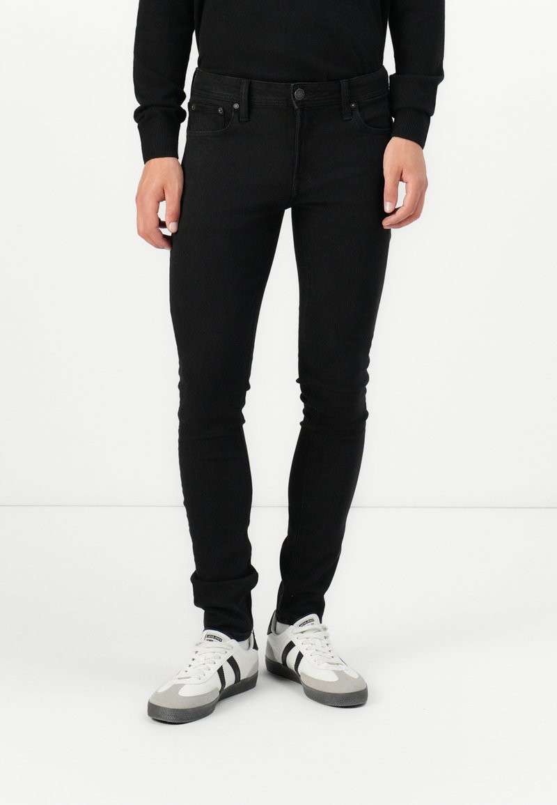 Black denim jeans with a slim fit, five-pocket design, and button closure. Paired with gray and white sneakers featuring black stripes.