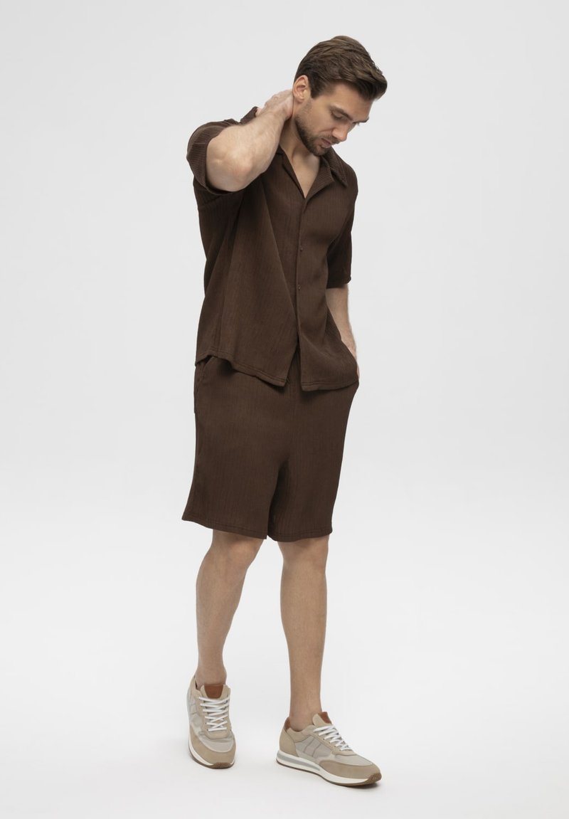 Homme portant une chemise marron à manches courtes boutonnée et un short assorti, debout avec une main sur la nuque et des baskets beiges.