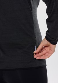Schwarzes, langärmliges Sportoberteil austexturiertem Material, mit seitlicher Reißverschlusstasche und körperbetonter Passform. Sanfter Stoff mit zartem Glanz.