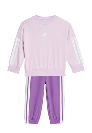 Lilaruskea collegepaita, jossa valkoiset raidat hihoissa, pyöreä kaulus ja painonapit. Yhteen sopivat violetit housut valkoisilla raidoilla sivuilla.