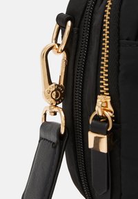 Guldmetallklämma som fäster svart läderrem vid svart väska med guldzipper och dragflik på vit bakgrund.