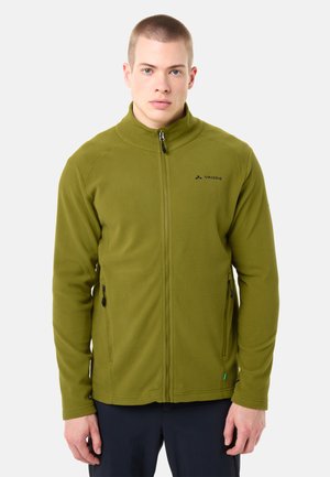 Jonge man met een olijfgroen fleecejack met ritssluiting en VAUDE-logo, staand tegen een effen lichte achtergrond, naar voren kijkend.