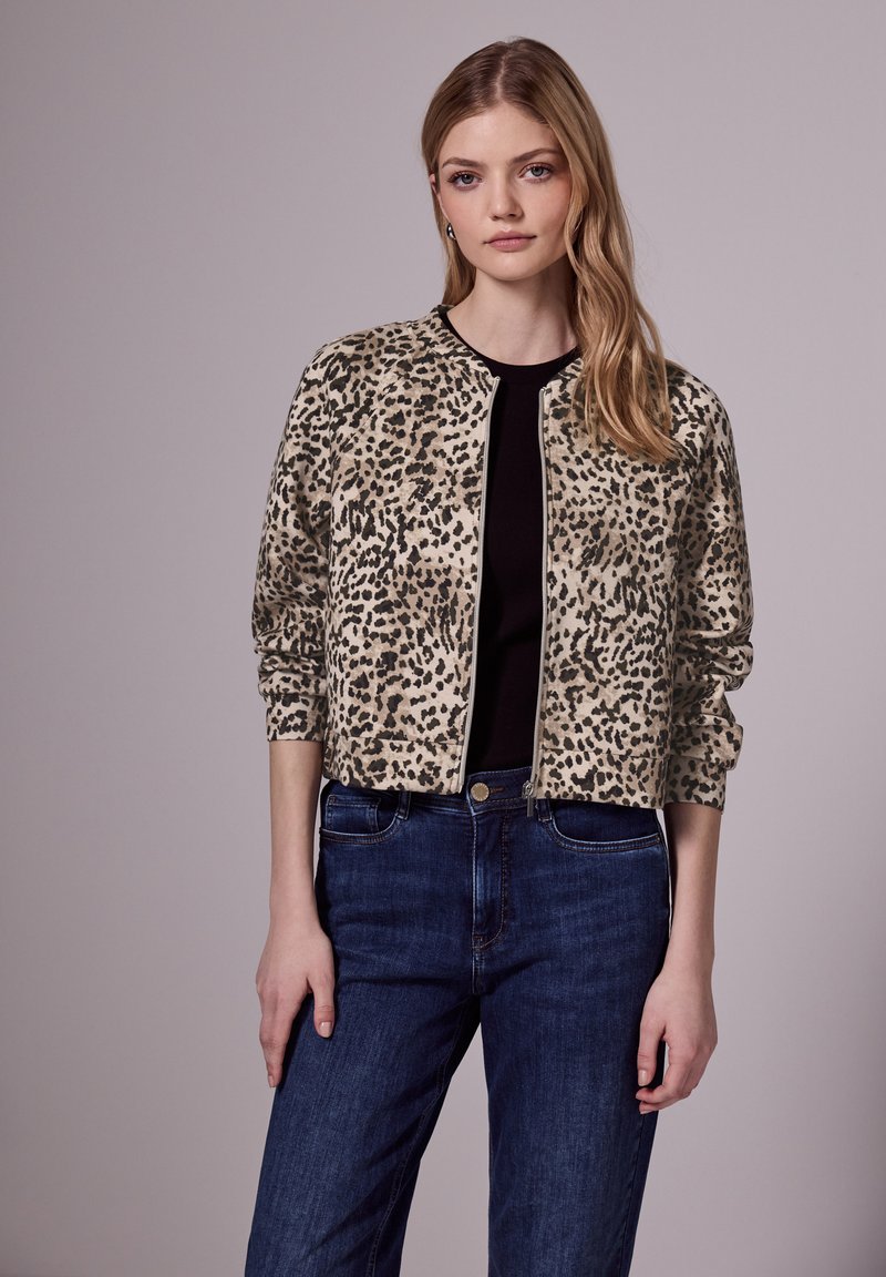 Bombers en imprimé léopard beige et noir, avec une fermeture éclair à l'avant, des poignets côtelés et une coupe décontractée, associés à un jean bleu foncé.