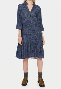 Vestito blu con motivo a disegni, scollo a V, maniche a tre quarti e orlo a balze. Abbinato a scarpe marroni e calzini grigi.