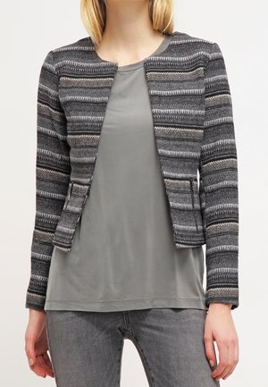 Vrouw draagt een aansluitende, open voorkant, gestreepte grijze en zwarte gebreide jas over een effen grijze blouse en grijze spijkerbroek, staand tegen een lichte achtergrond.