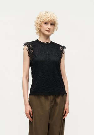 Γυναίκα με κοντά σγουρά ξανθά μαλλιά, φορώντας μαύρο δαντελένιο αμάνικο τοπ και άνετο παντελόνι ελιάς, στέκεται μπροστά σε απλό φόντο.