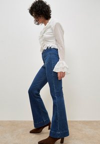 Blusa bianca con volant e jeans a vita alta blu svasati. Stivaletti marroni a punta. Design semplice e testurizzato con contrasto di colori chiari e scuri.