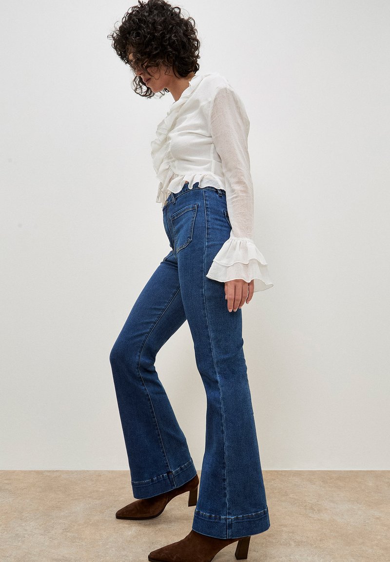 Blusa bianca con volant e jeans a vita alta blu svasati. Stivaletti marroni a punta. Design semplice e testurizzato con contrasto di colori chiari e scuri.