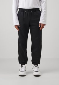 Pantalones de chándal negros con cintura elástica y cordón ajustable. Tela suave, piernas entalladas y tobillos acampanados. Combinados con zapatillas blancas.