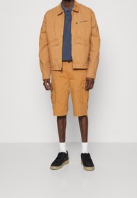 Hellbraune Baumwollbomberjacke mit Reißverschlusstasche, kombiniert mit hellbraunen Cargo-Shorts. Das Model trägt ein navyblaues Polohemd und schwarze Sneakers mit weißen Socken.