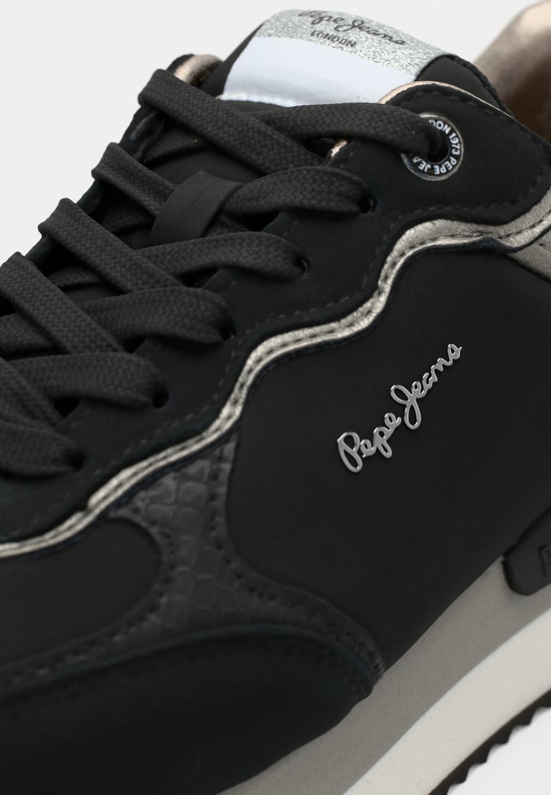 Schwarzer Sneaker mit strukturierten Lederakzenten, flachen Schnürsenkeln und silbernem Logo. Verfügt über eine hellfarbige Sohle und eine glänzende Zunge mit Markenlogo.