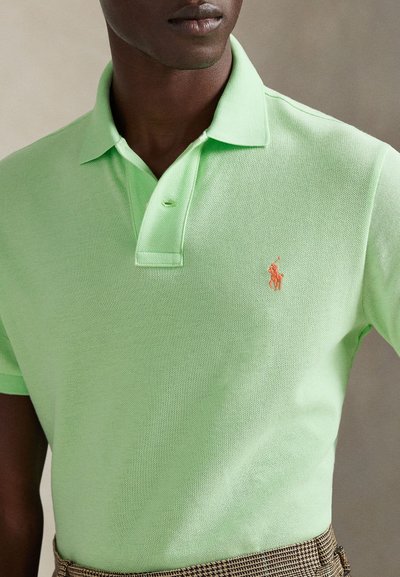 Ανοιχτό πράσινο πόλο πουκάμισο από υφή υφάσματος, με πλάκα δύο κουμπιών και μικρό πορτοκαλί κεντημένο λογότυπο στο αριστερό στήθος.