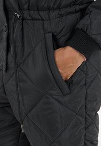 Zwarte gewatteerde jumpsuit met een elastische tailleband, contrasterende geribde manchetten en zijzakken. Het materiaal lijkt soepel en geïsoleerd.