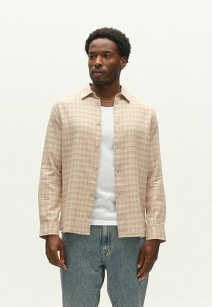 Mann trägt beige kariertes Hemd mit Knöpfen über weißem Tanktop und blauen Jeans, steht vor einem schlichten, hellen Hintergrund.