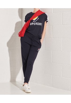 Camiseta de algodón azul oscuro con el logo de "Superdry" y acentos rojos, combinada con joggers oscuros a juego y zapatillas blancas. Sudadera roja colgada.