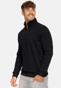 Sweat à col zippé noir en tissu doux, doté de manches longues, de poignets et d'ourlet côtelés, avec un petit logo sur la poitrine.