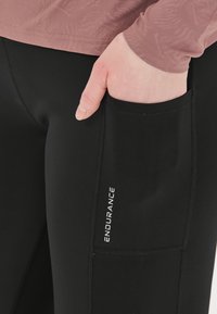 Musta urheiluleggingsit, joissa on sivutasku, sileällä pinnalla ja valkoisella "ENDURANCE" -brändäyksellä reidessä.